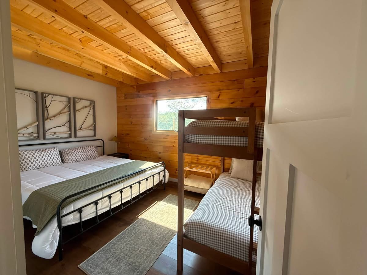 Rental cabin bunk room with a made king bed, white bedding, and gingham-covered bunk beds under a wood beam ceiling