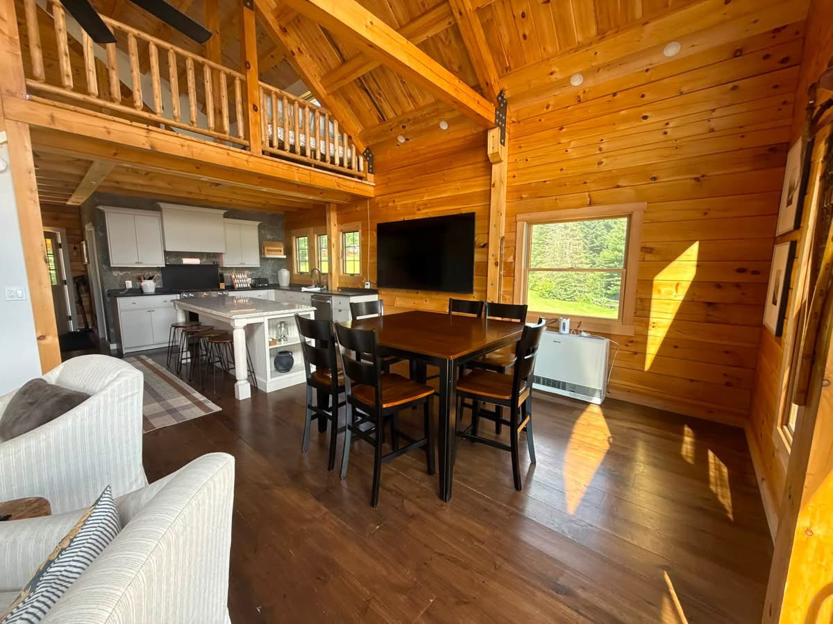 Sunlit log cabin great room with an open white kitchen, dining table, and a loft railing overhead, freshly cleaned